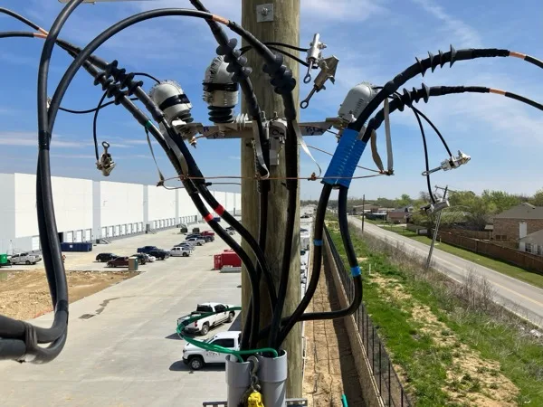Medium voltage riser terminations at the top of a pole overlooking a white tilt wall distribution center and its truck court