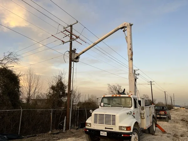 Bucket truck parked beneath a distribution pole line at dusk beside a fenced yard