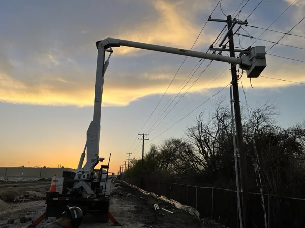 Bucket truck boom raised to a riser pole at sunset with an industrial building in the background