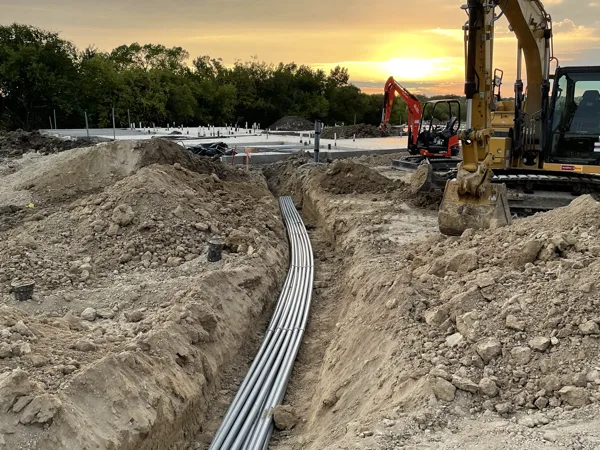 Excavator beside a trench with a bundle of conduit laid in it at sunset