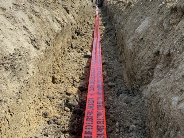 Open trench with buried electric line caution tape and an excavator behind
