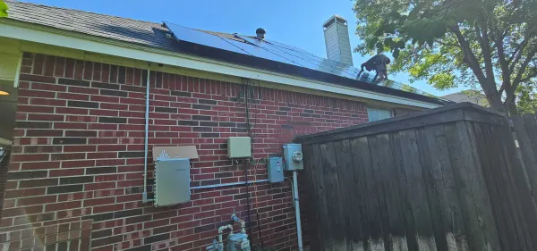R&B Power installer securing modules on a shingle roof in Grapevine, Texas, with the Enphase combiner and disconnect mounted on the brick wall below